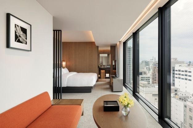 Interior view of a modern hotel room featuring a comfortable bed, a stylish orange sofa, and large windows with a cityscape view.