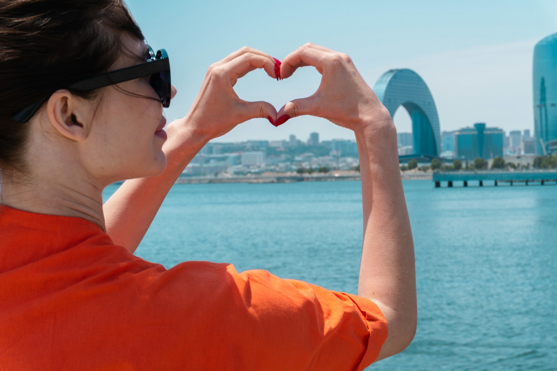 A woman in an orange top makes a heart with her hands, posed near the water in a city